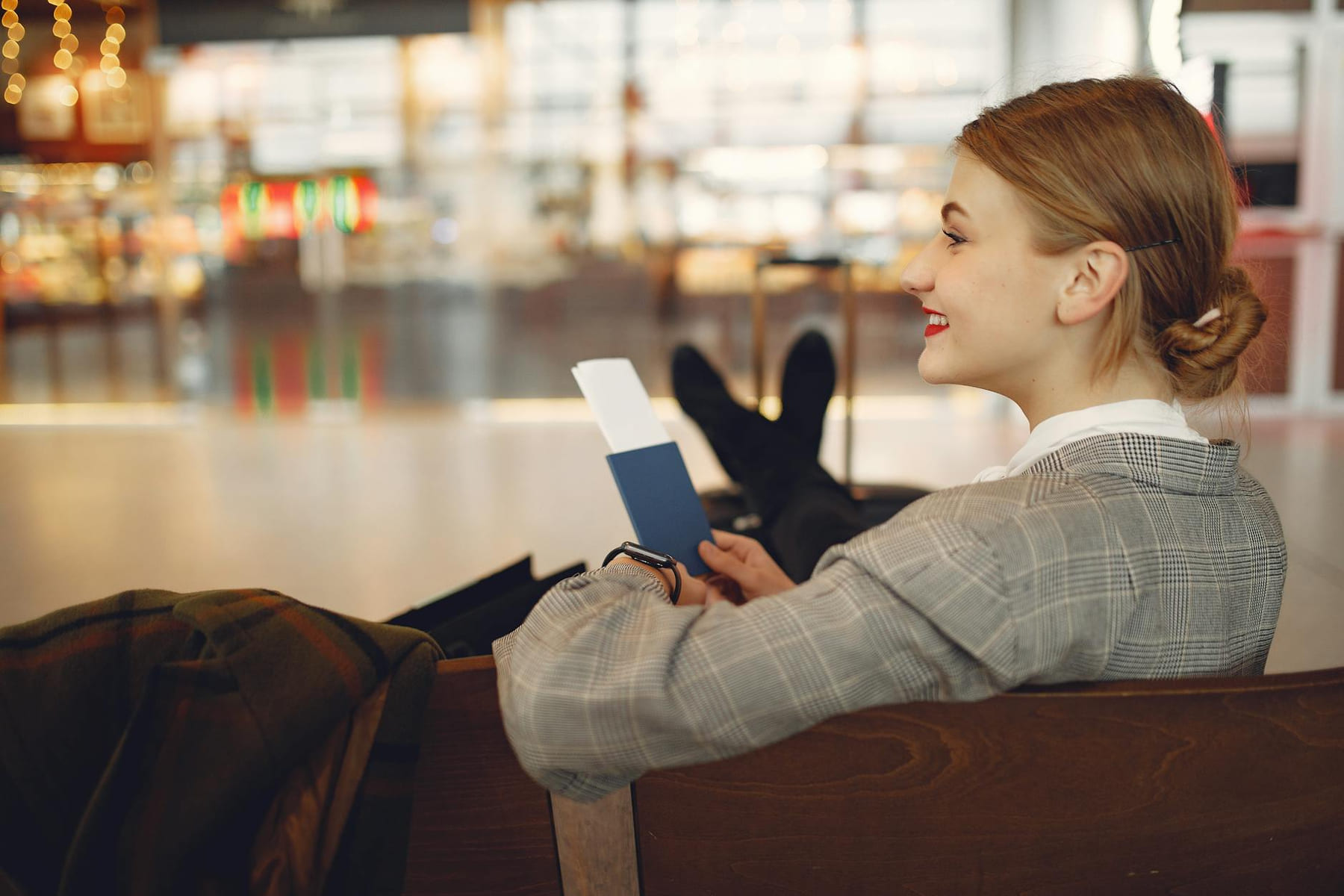 Side view of cheerful female student in checkered jacket smiling away while chilling in hall with outstretched legs and passport in hands on Points and Pathways